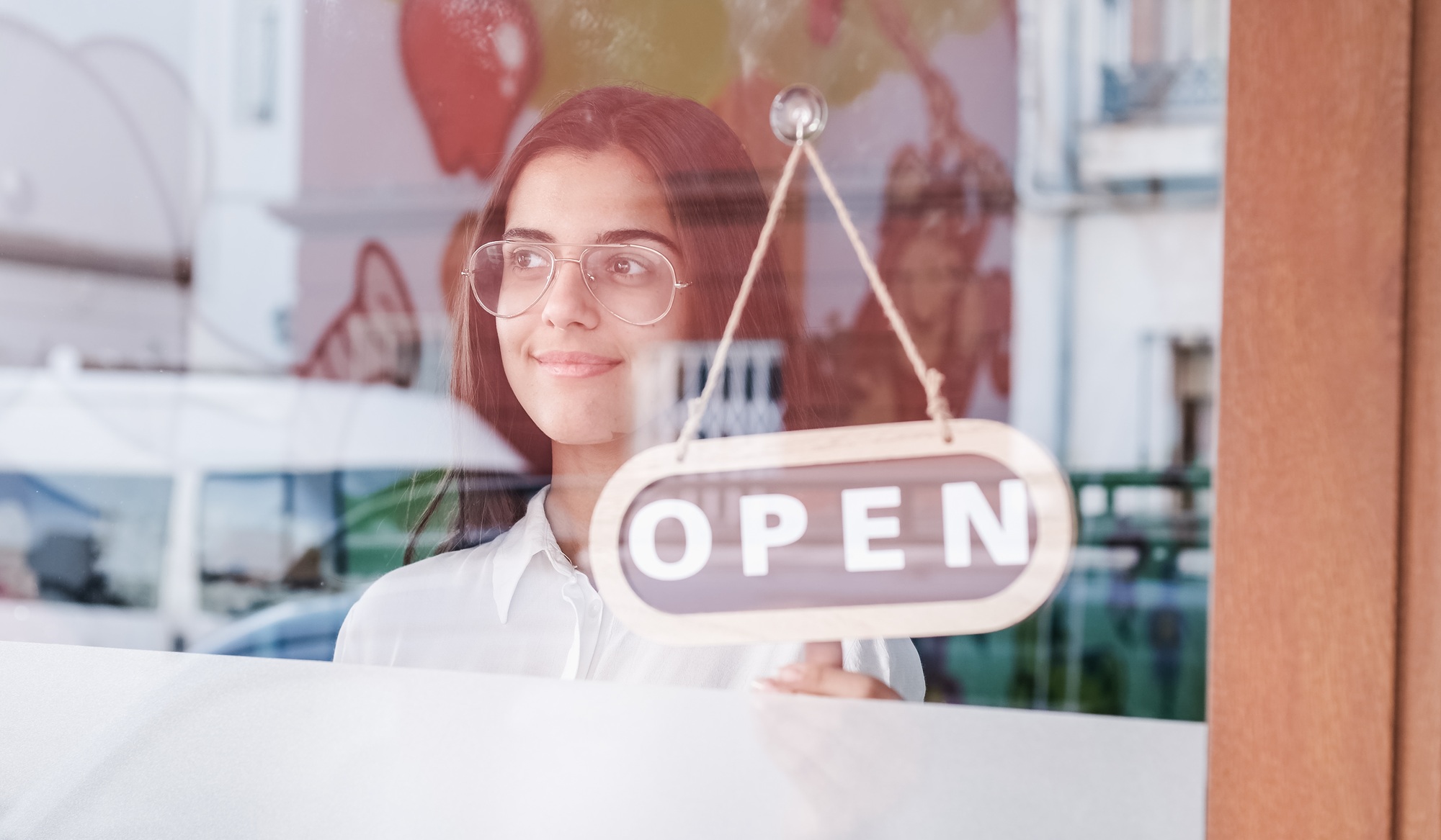 Happy small business girl changing closed to open sign on window smiling looking outside